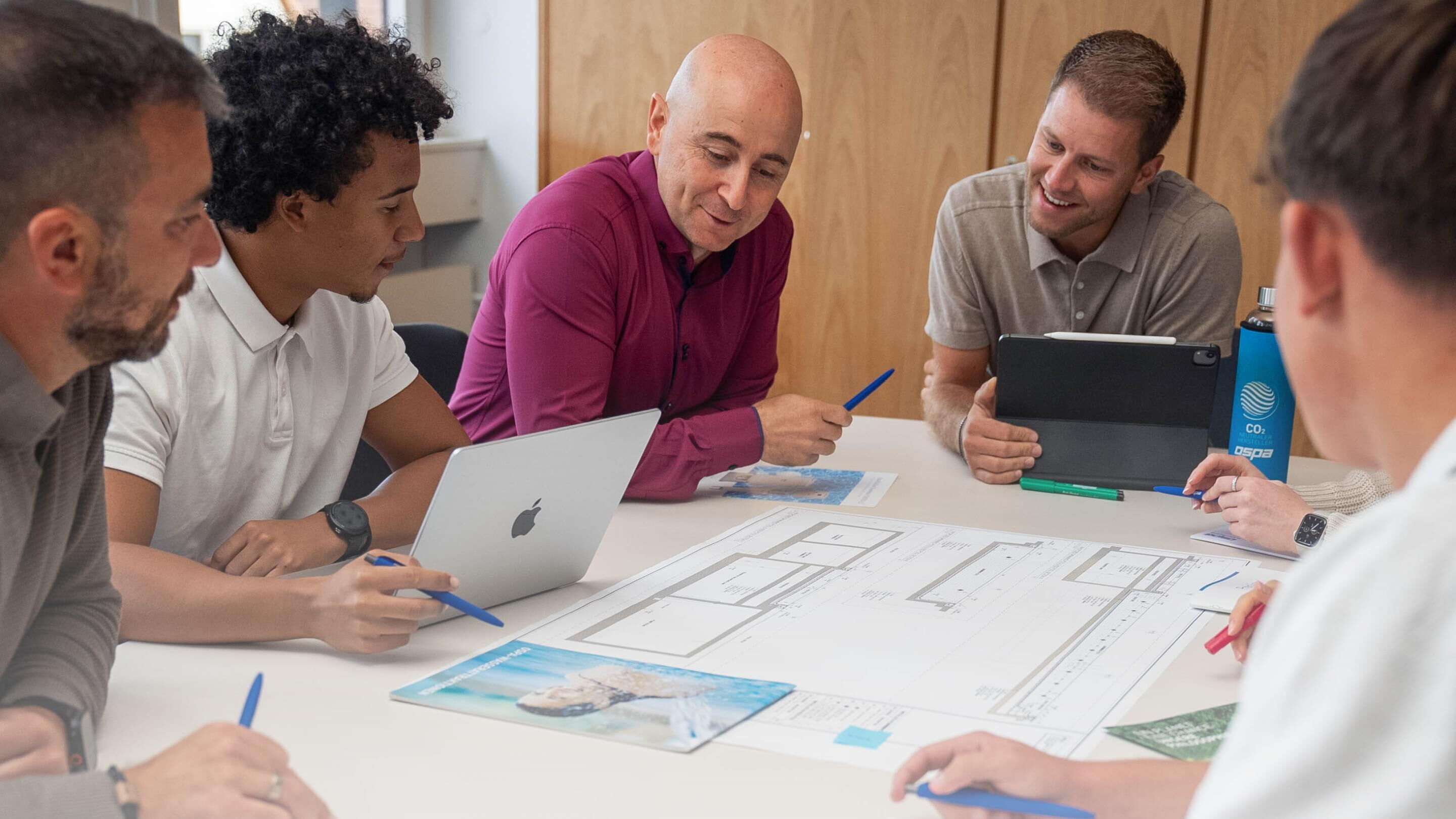 Ein lächelnder Mann steht an einem Tisch, während sein Team an einem Laptop und Projekten arbeitet.