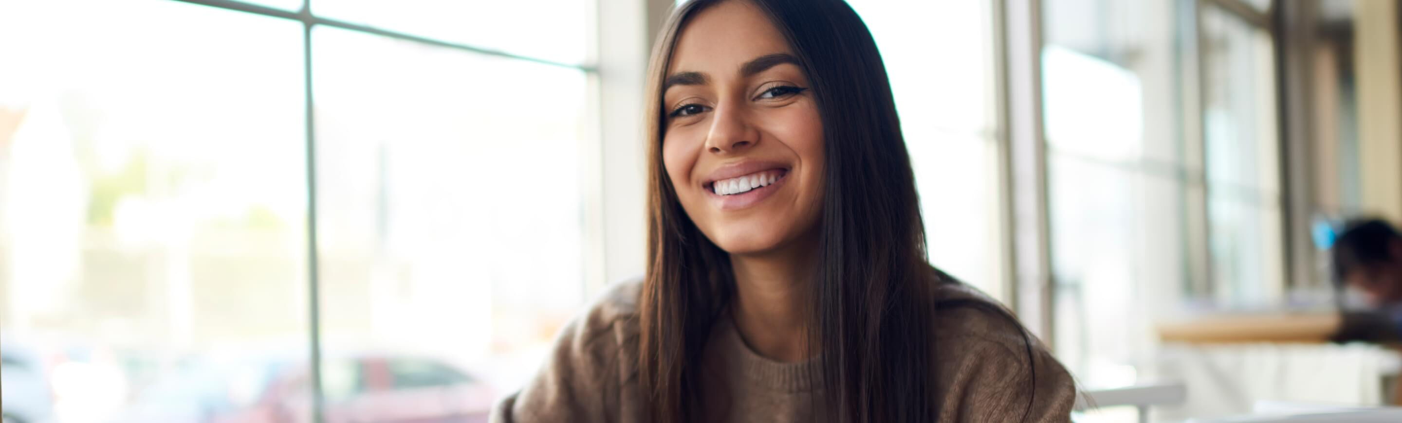 Eine junge Frau mit langen Haaren lächelt fröhlich in einem modernen Café, während sie am Tisch sitzt.