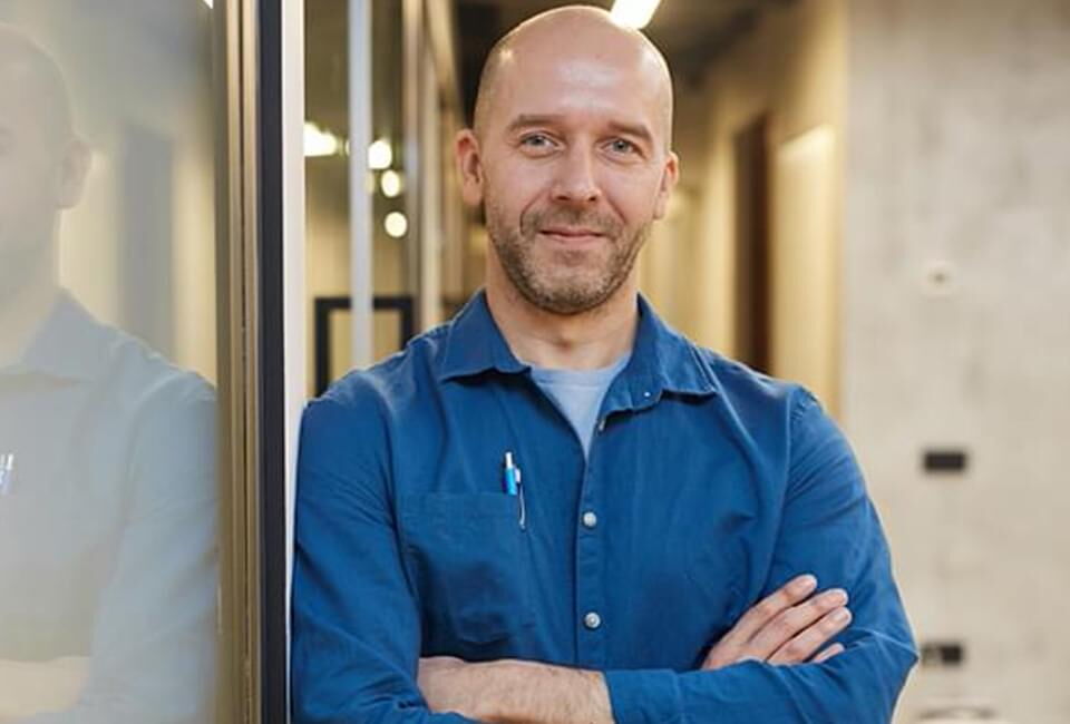 Ein lächelnder Mann in einem blauen Hemd steht in einem modernen Büro mit verschränkten Armen.