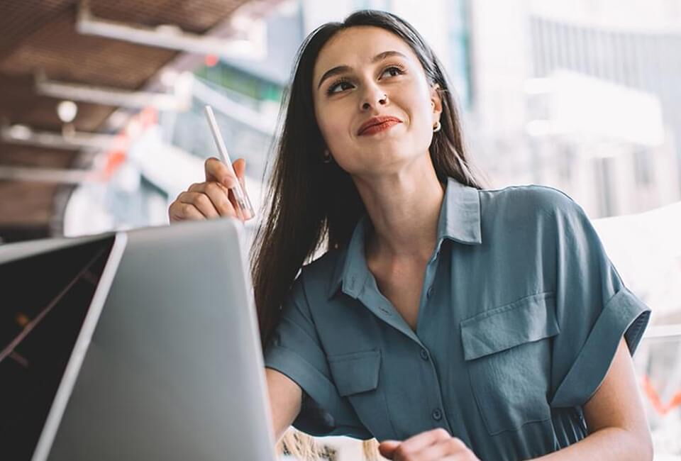 Eine junge Frau mit einem Stift in der Hand lächelt nachdenklich am Laptop in einem modernen Büro.
