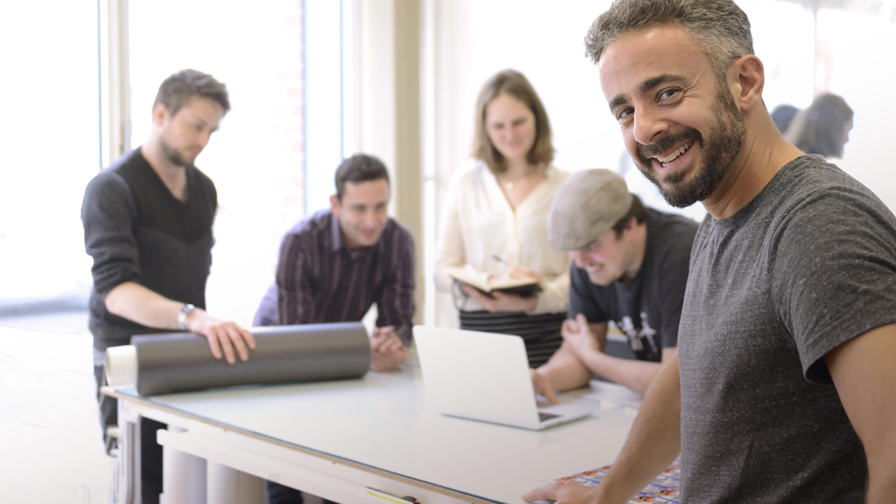 Ein lächelnder Mann steht an einem Tisch, während sein Team an einem Laptop und Projekten arbeitet.