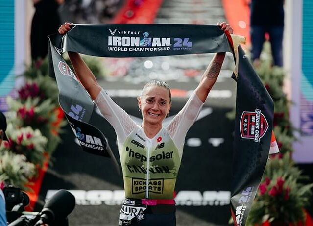 Das Bild zeigt Laura Philipp, wie sie die Ziellinie der Ironman-Weltmeisterschaft 2024 überquert. Sie lächelt und hält ein Banner hoch, auf dem “VinFast Ironman 24 World Championship” steht. In einem Rennanzug mit den Logos “hepsolar” und “Kickass Sports”